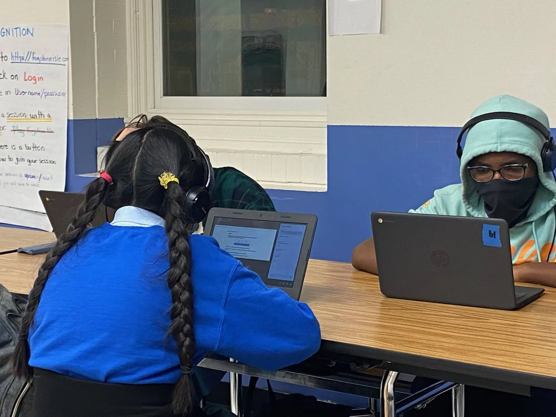 Two masked students working on laptops at a table