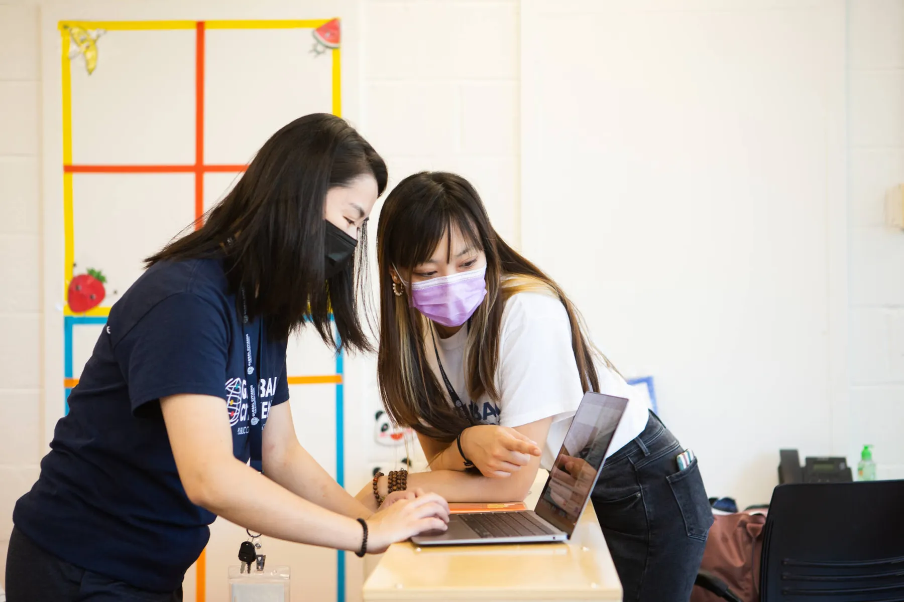 Two masked women leaning over a laptop at a classroom desk