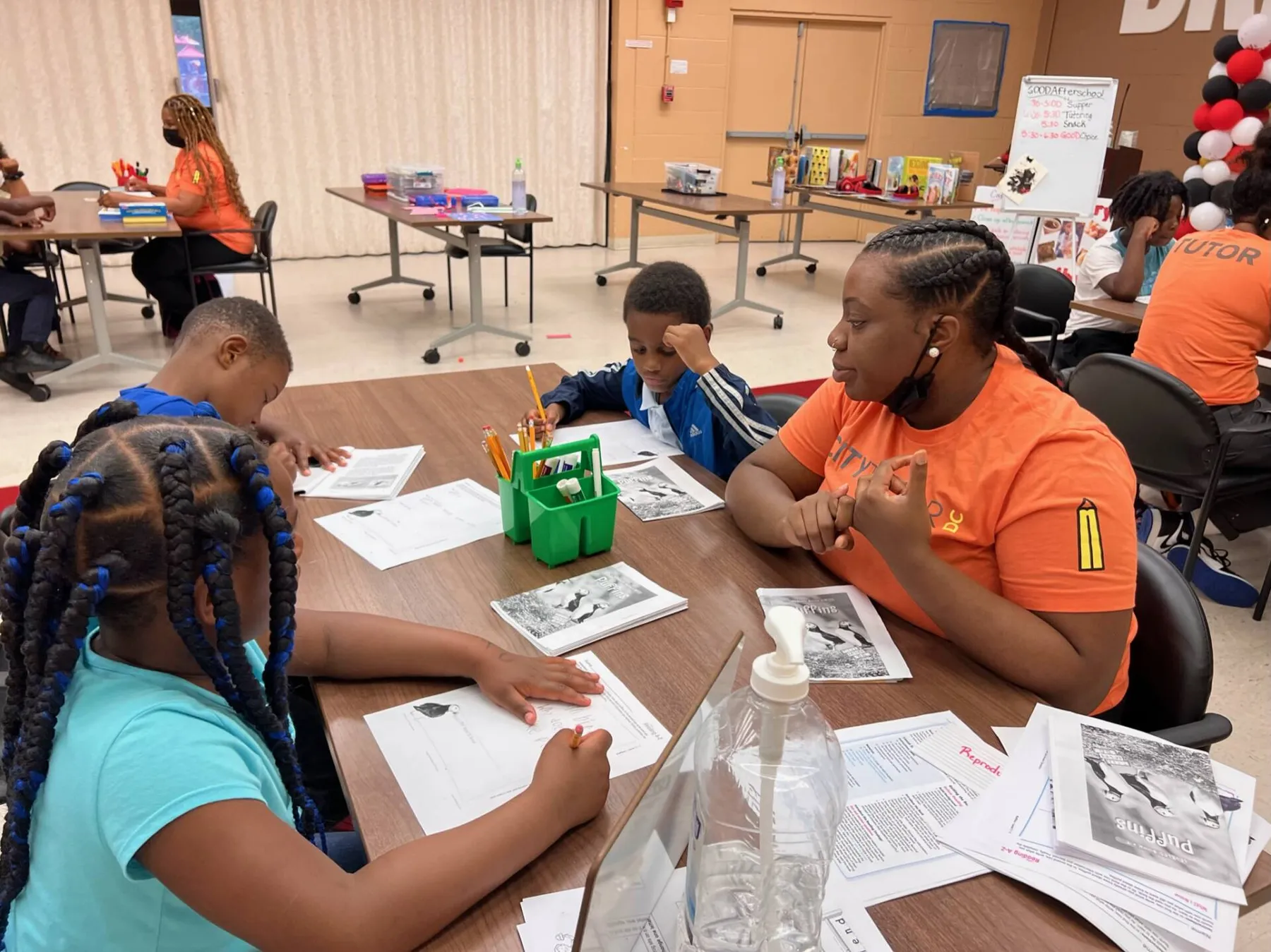 Tutor in an orange shirt leading three children working with worksheets at a table
