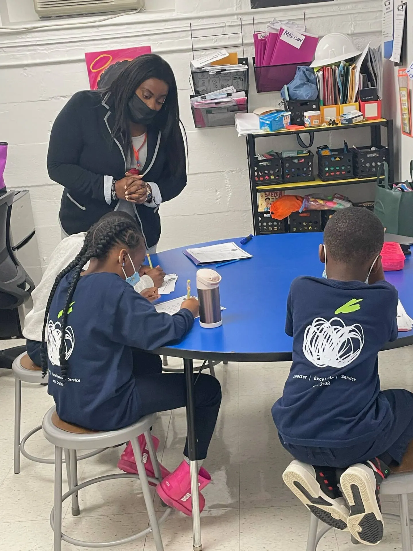 Woman standing beside three children seated around a blue classroom table