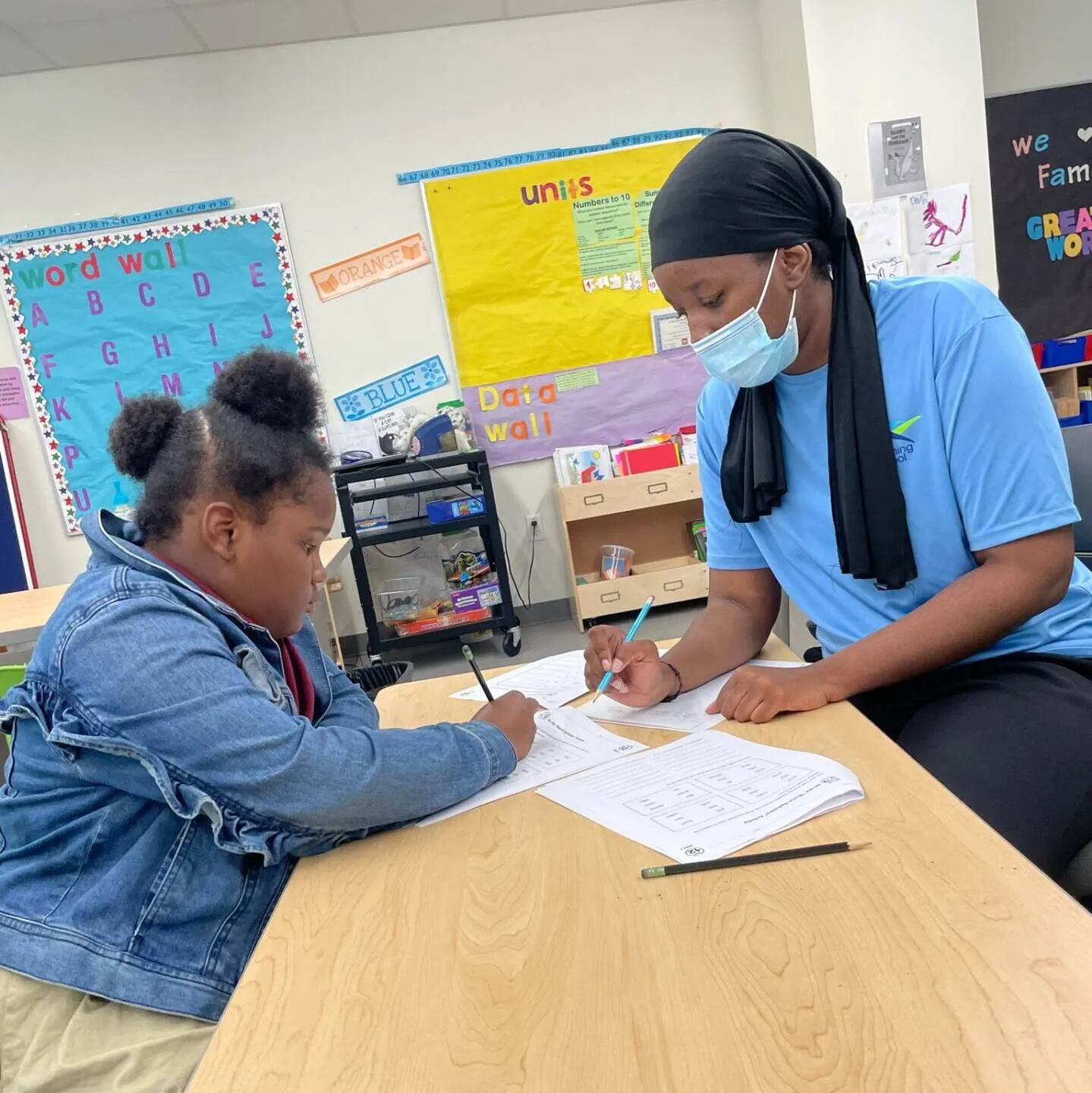 Masked woman helping a seated child complete a worksheet