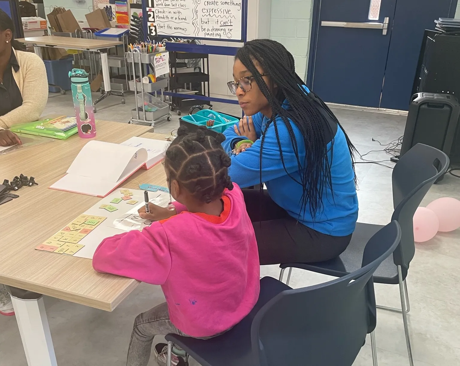 Woman in a blue shirt helping a girl in a pink shirt with papers at a table