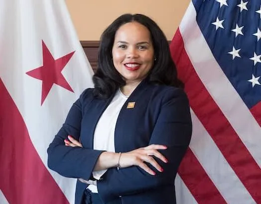 Woman in a navy suit posing with folded arms between DC and United States flags