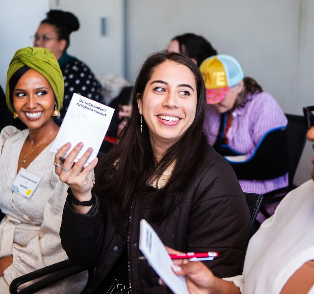 Two women seated at an event, one holding up a printed card