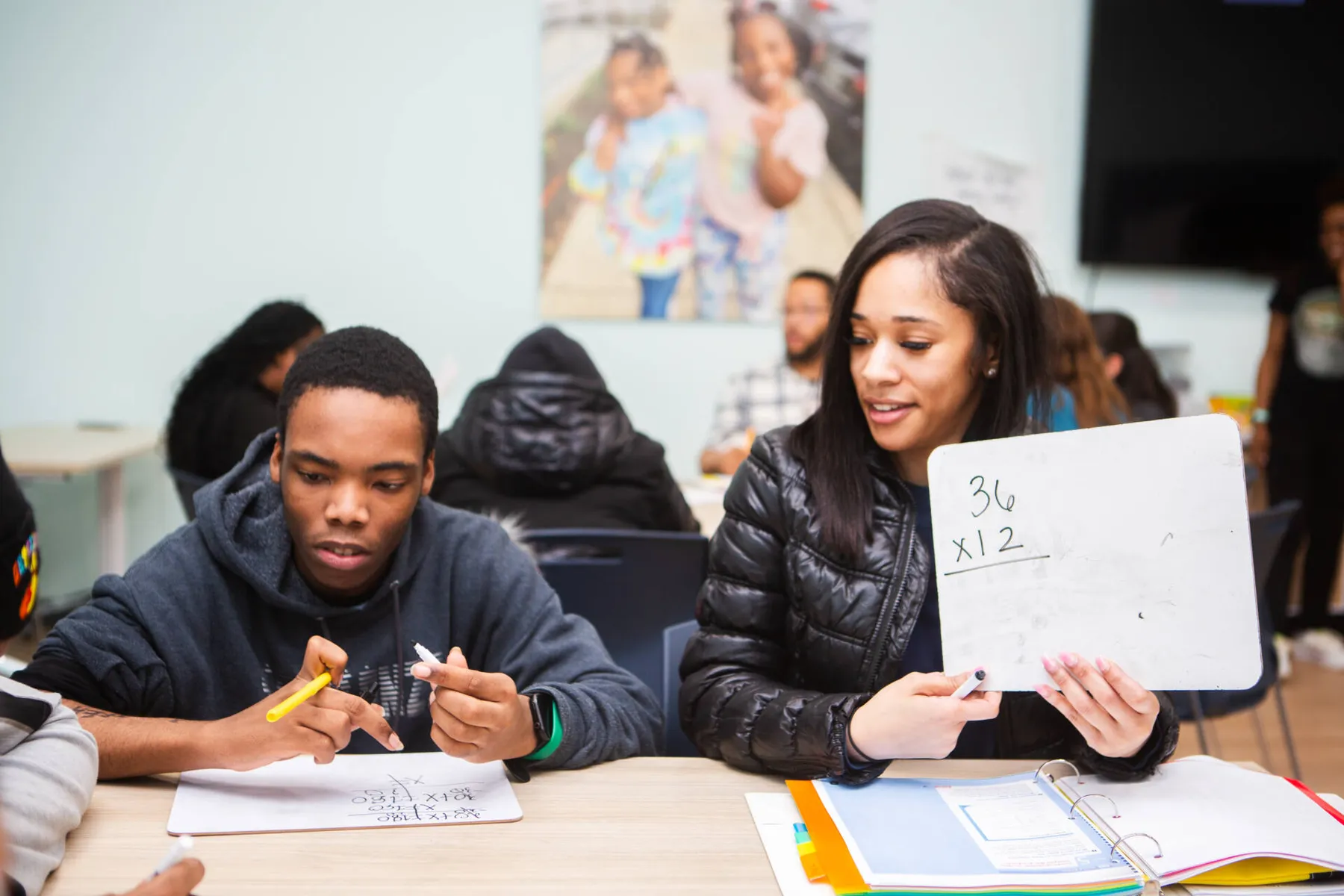 Two students at a table while one holds up a handwritten math page