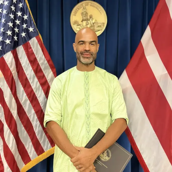 Man in a pale green shirt posing between United States and District of Columbia flags