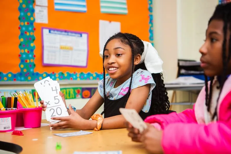 Girl holding up a handwritten math answer at a classroom table
