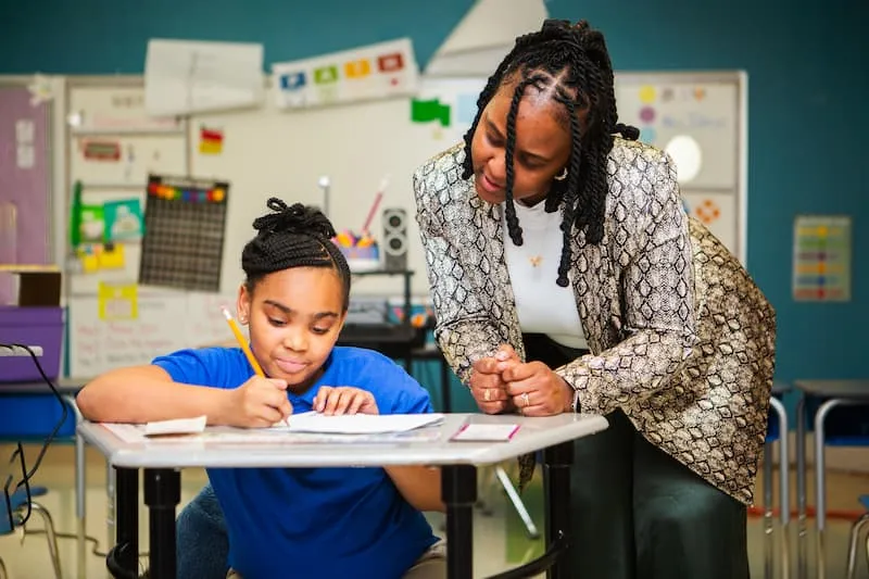 Woman leaning over a seated student and pointing at a worksheet